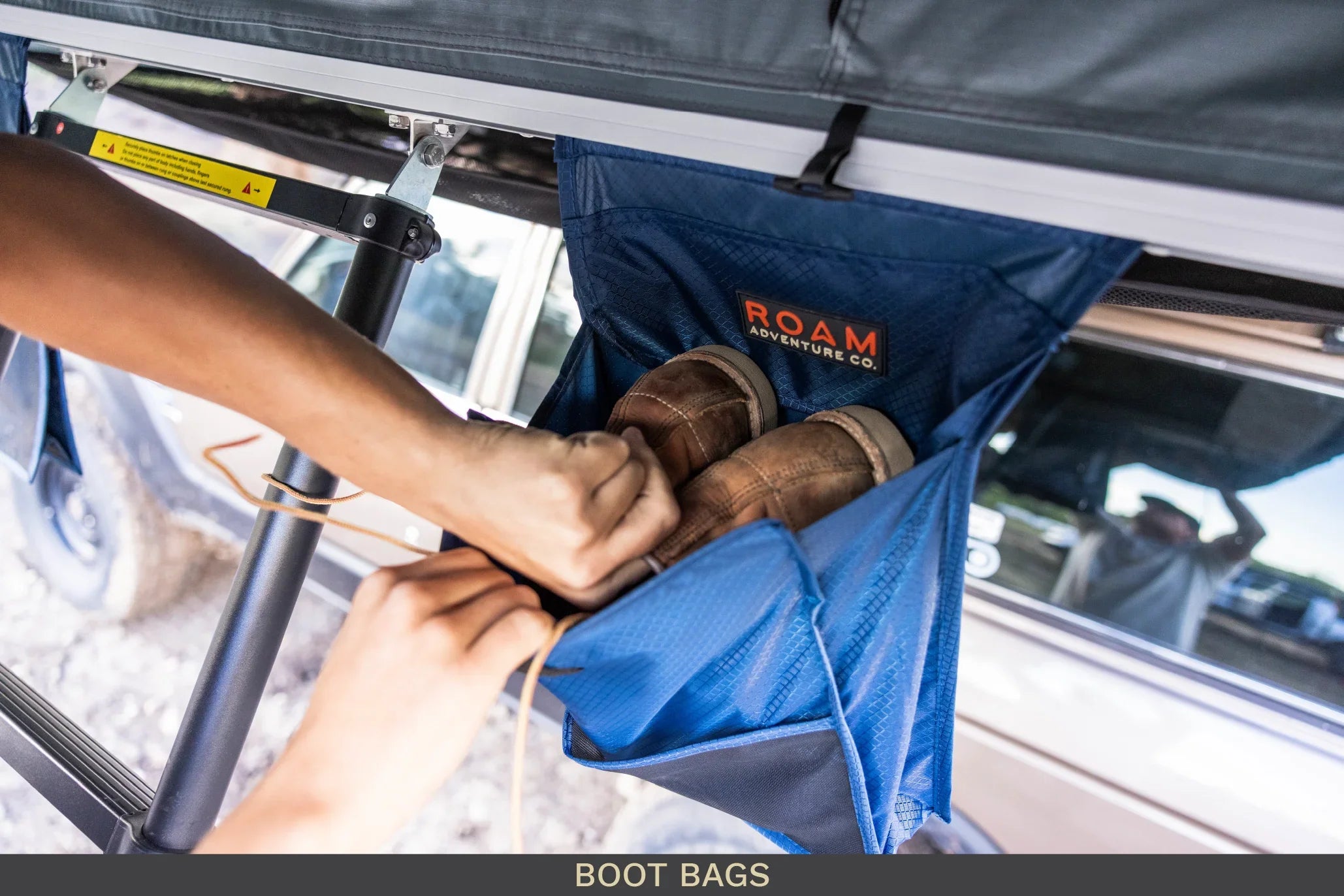 Hands placing boots into a blue overland camping boot bag attached to a vehicle rack