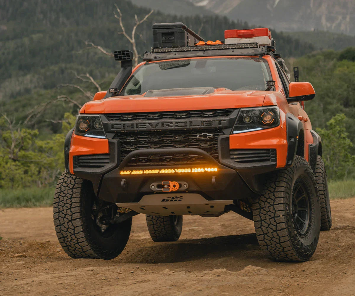 Orange Chevrolet off-road truck with roof rack, gear, and LED light bar on dirt trail